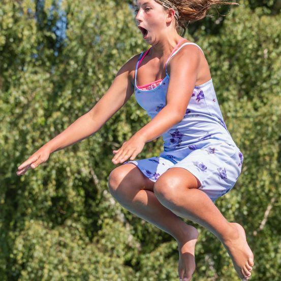 Ein junges Mädchen in der Luft mit offenem Mund, trägt ein blaues Blumenkleid und springt von einer Sprungbrett mit grünem Hintergrund.