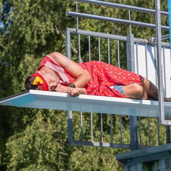Ein Mann in einem roten Outfit und einem gelben Bandana schläft auf einer Sprungbrett mit einem Metallgeländer im Hintergrund.