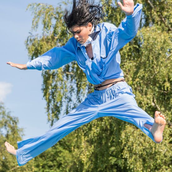 Eine Frau in einem blauen Overall springt in die Luft, mit erhobenen Händen, während ihr Haar im Wind weht, vor einem Hintergrund aus grünen Bäumen und blauem Himmel.