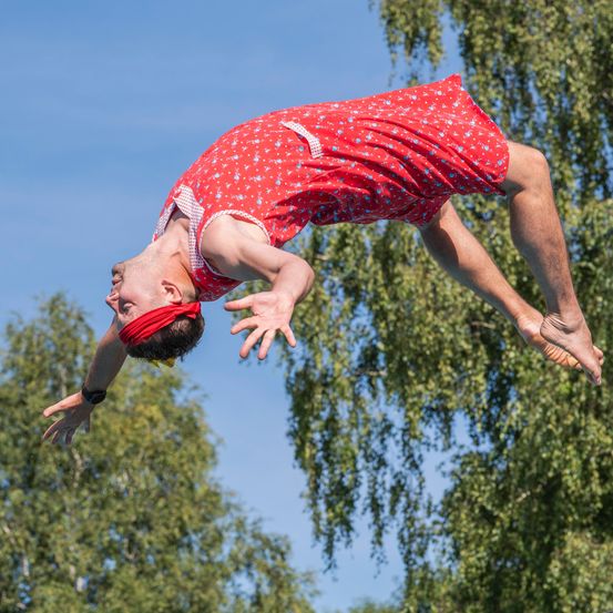 Ein Mann ist in der Luft, auf dem Kopf stehend, trägt ein rotes Blumenkleid und eine rote Kopfband. Er führt einen akrobatischen Trick vor. Der Himmel ist blau.