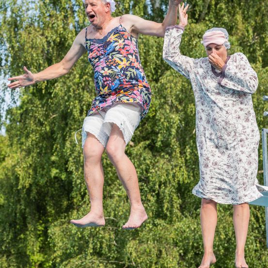 Ein älterer Mann und eine ältere Frau springen von einer Sprungturm, der Mann trägt ein Blumen-Tanktop und die Frau ein Blumenkleid, beide tragen Mützen.