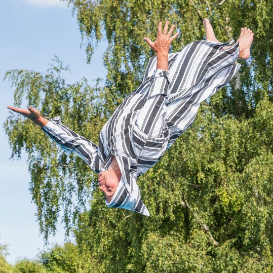 Eine Person in gestreiften Pyjamas schwebt auf dem Kopf, mit ausgebreiteten Armen und Beinen, vor dem Hintergrund von grünen Bäumen und blauem Himmel.