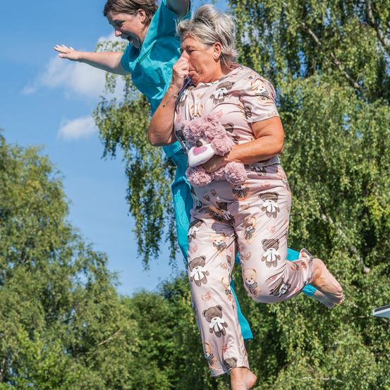 Zwei Frauen springen in der Luft; eine trägt einen Schlafanzug mit einem Teddybären, die andere ein blaues Outfit und hält die Füße der ersten Frau. Sie sind draußen bei klarem Himmel.