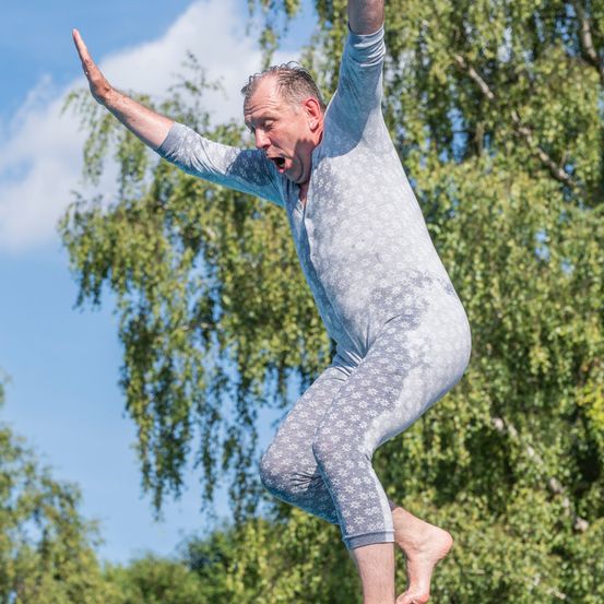 Ein älterer Mann springt mit offenem Mund und erhobenen Armen vor einem Baum in der Luft.
