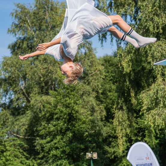 Ein Mädchen macht einen Handstand auf einer Sprungbrett, die Beine in der Luft, mit einem weißen Kleid und Socken. Im Hintergrund sind grüne Bäume und ein Banner mit weißem Text zu sehen.