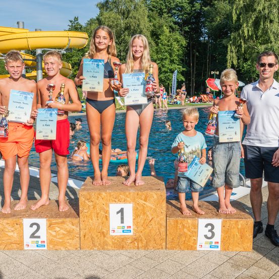 Fünf junge Personen stehen auf einem Podium und halten Trophäen und Urkunden. Zwei Mädchen in Badekleidung stehen auf dem mittleren Podium, mit der Nummer eins darauf. Drei Jungen stehen auf den anderen Podesten, jeder hält eine Trophäe. Der Hintergrund zeigt einen Pool mit einer Wasserrutsche.