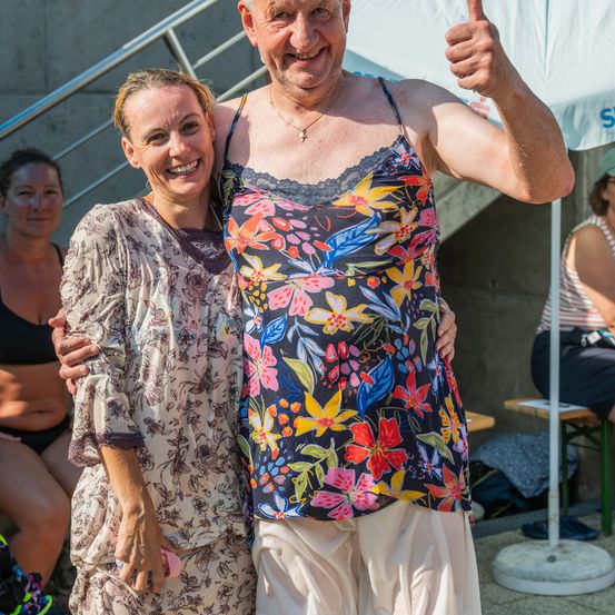 Ein älterer Mann und eine Frau stehen zusammen und lächeln für ein Foto. Der Mann zeigt den Daumen nach oben. Sie stehen in der Nähe einer Treppe mit Geländer. Hinter ihnen sitzt eine weitere Frau auf einer Bank.