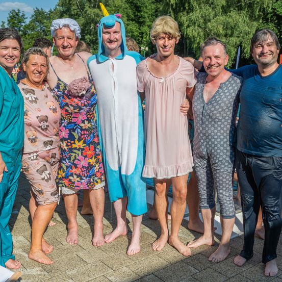 Eine Gruppe von Erwachsenen, in verschiedenen Kostümen gekleidet, steht zusammen auf einem Außengelände. Ein Mann trägt ein Einhorn-Onesie, ein anderer ein rosa Nachthemd. Dahinter sind Bäume und ein Pool zu sehen.
