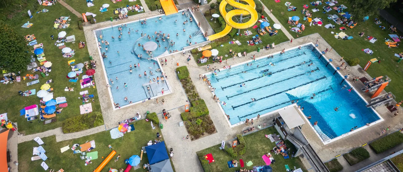 Eine Luftaufnahme eines großen Freibadkomplexes mit zwei Schwimmbecken, einer gelben Wasserrutsche und vielen Menschen, die den Tag genießen, einige schwimmen, andere sonnenbaden und wieder andere faulenzen.