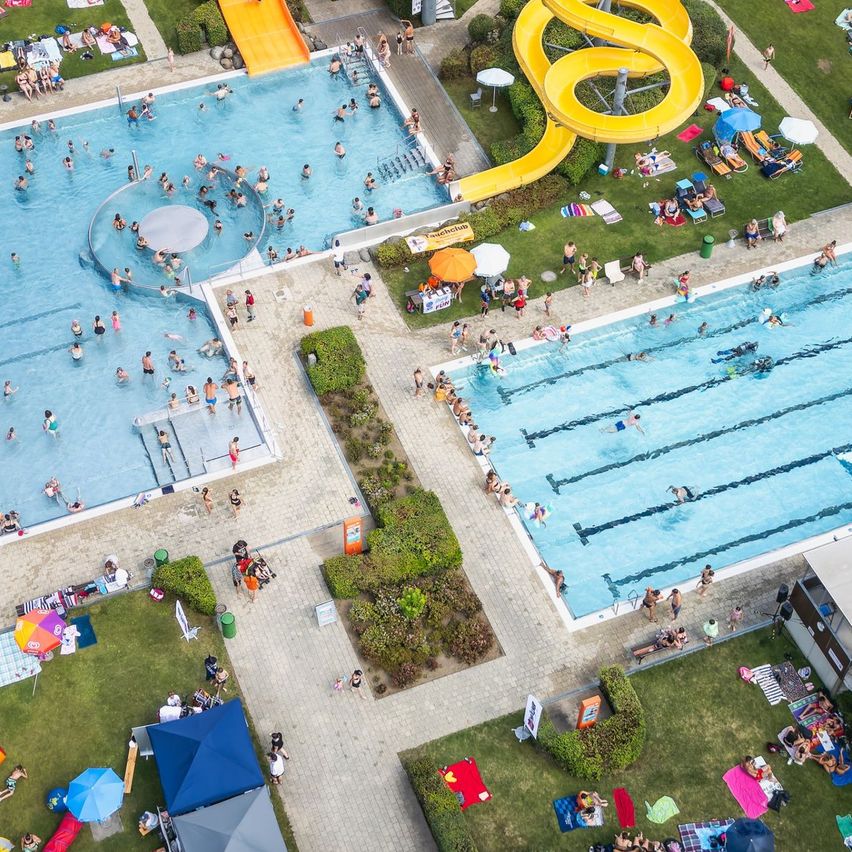 Eine Luftaufnahme eines großen Freibadkomplexes mit zwei Schwimmbecken, einer gelben Wasserrutsche und vielen Menschen, die den Tag genießen, einige schwimmen, andere sonnenbaden und wieder andere faulenzen.