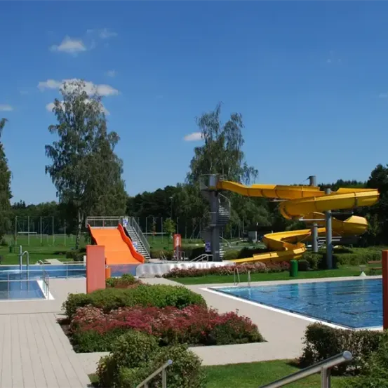 Freibad mit zwei Wasserrutschen und einer großen Terrasse, umgeben von Grünflächen und Bäumen.
