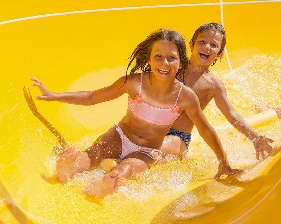 Zwei Kinder rutschen einen gelben Wasserrutschen hinunter, beide lächeln und genießen die Fahrt. Das Mädchen trägt einen rosa Bikini, während der Junge oberkörperfrei ist.