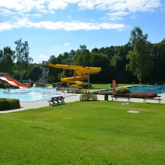 Ein Freibad mit zwei Rutschen, eine gelb und eine rot, unter einem blauen Himmel mit einigen Wolken. Bäume und Büsche umgeben das Gebiet.