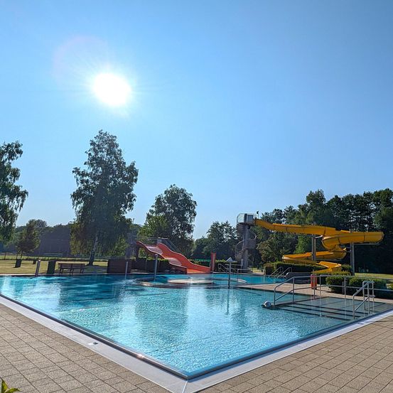 Ein Freibad mit zwei Rutschen, eine rot und eine gelb, unter einem klaren blauen Himmel mit strahlender Sonne.