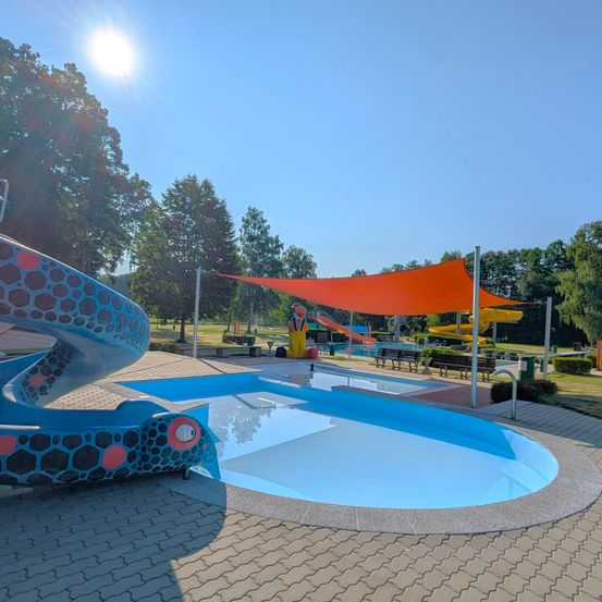 Freibad mit einer Rutsche und einem orangefarbenen Sonnensegel, unter einem sonnigen Himmel. Bäume und Grünflächen im Hintergrund.