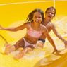 Two children, a girl and a boy, are sliding down a yellow water slide, smiling and holding onto each other for support.