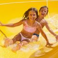 Zwei Kinder, ein Mädchen und ein Junge, rutschen einen gelben Wasserrutsche hinunter, lächeln und halten sich gegenseitig an, um Halt zu haben.