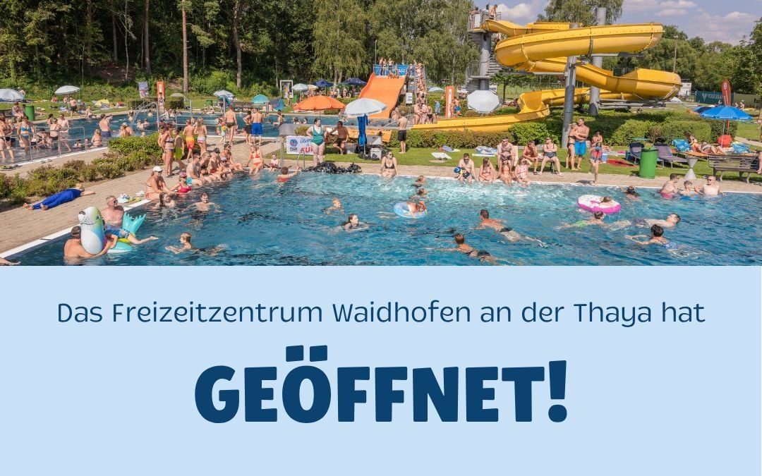 A large outdoor water park with multiple pools and water slides. Many people are swimming and enjoying the water. The park is surrounded by trees and has a clear blue sky.