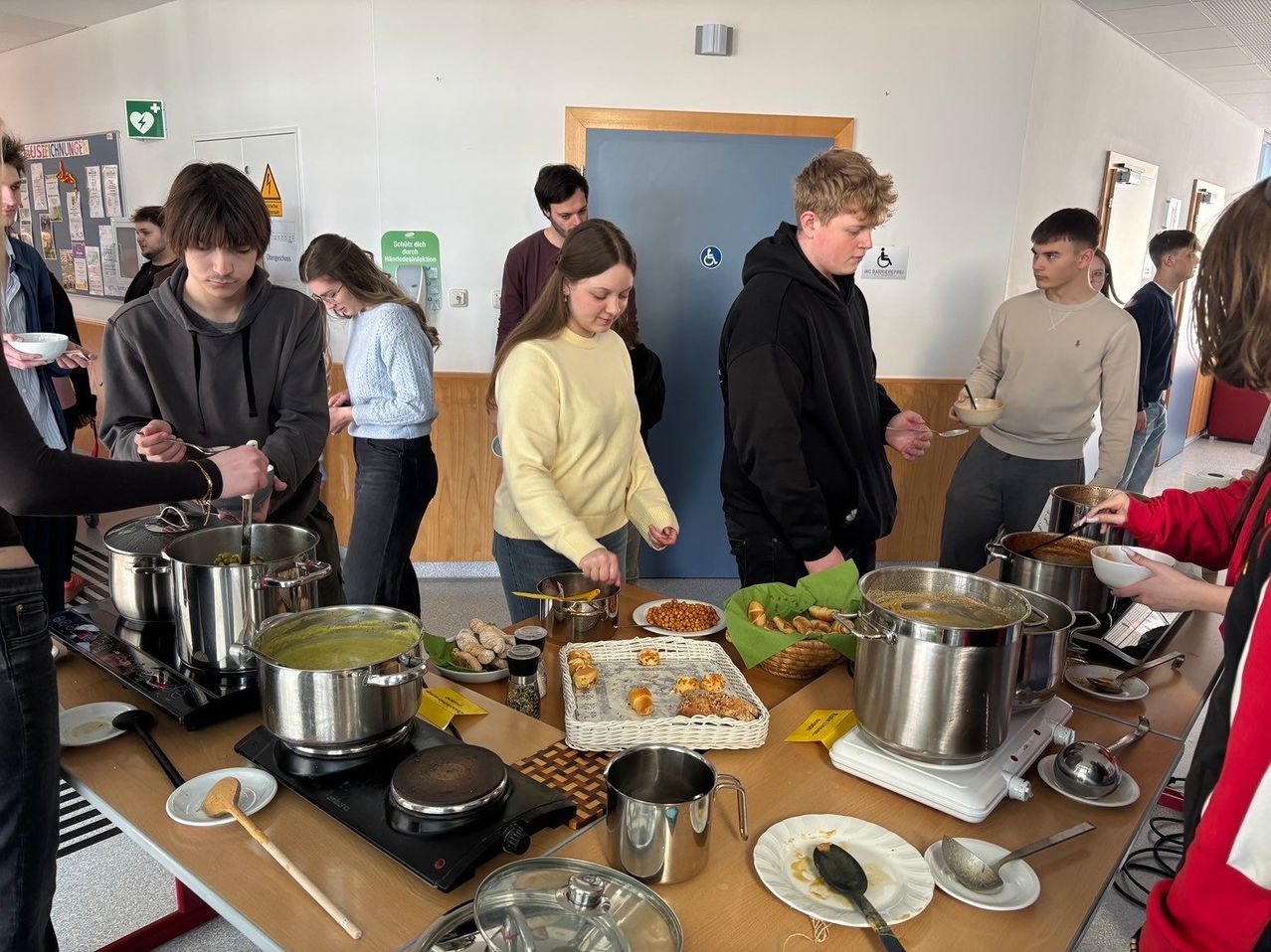 A group of people are gathered around a table with various food items and pots. Some are cooking, while others are eating. They seem to be participating in a cooking event or a meal preparation.