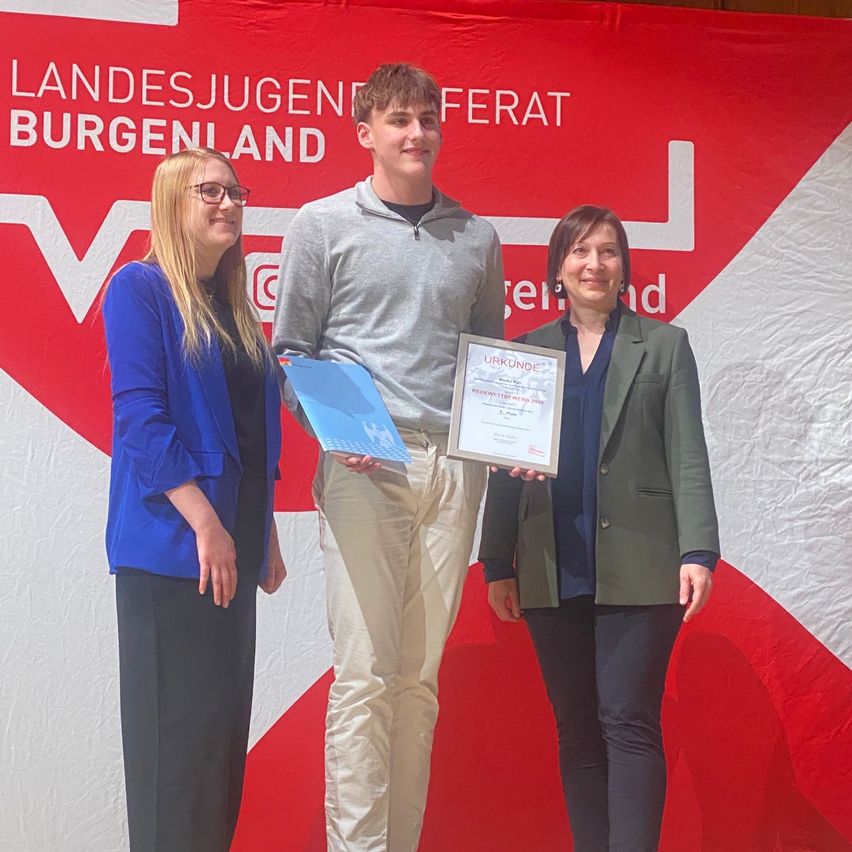 Three people stand on stage. A young man holds a certificate, while two women smile, one holding a blue folder. The backdrop reads 'Landesjugendrat Burgenland'.