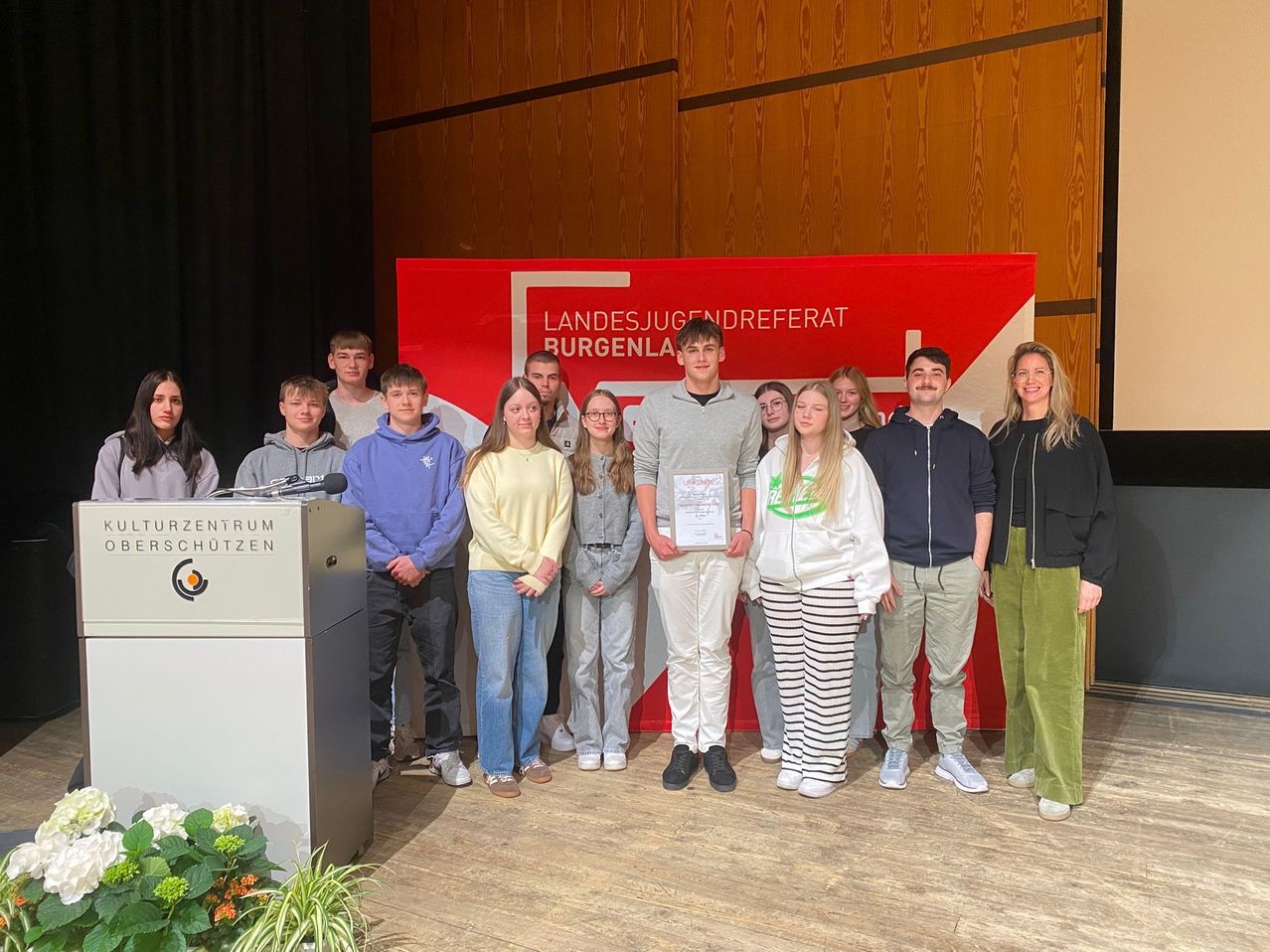 A group of young people stands on a stage, smiling. One person holds a certificate. Behind them is a red banner with the text Landesjugendreferat Burgenland.