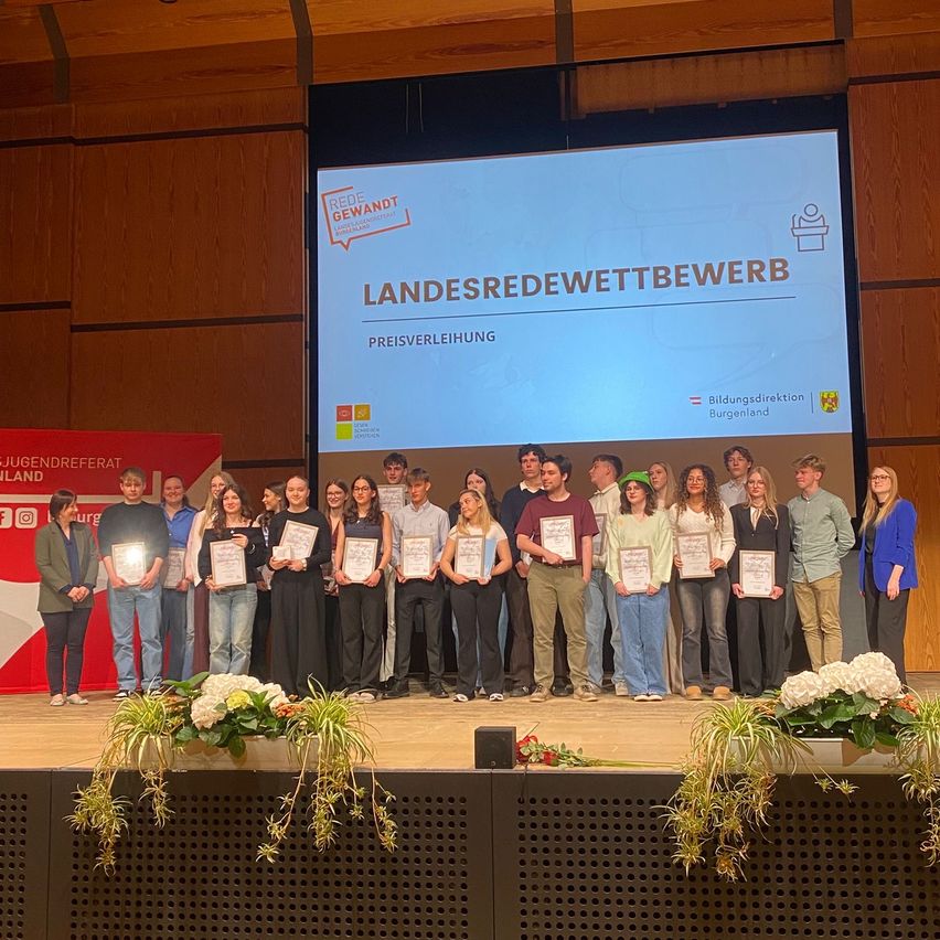 A group of people stand on a stage, holding certificates, with a screen behind them displaying 'Landesredenwettbewerb'. They are likely at an award ceremony.