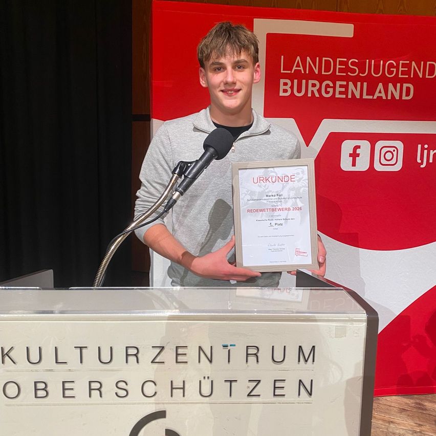 A young man stands at a podium holding a certificate with a red and white backdrop behind him. The backdrop reads Landesjugend Burgenland. He stands next to a microphone.