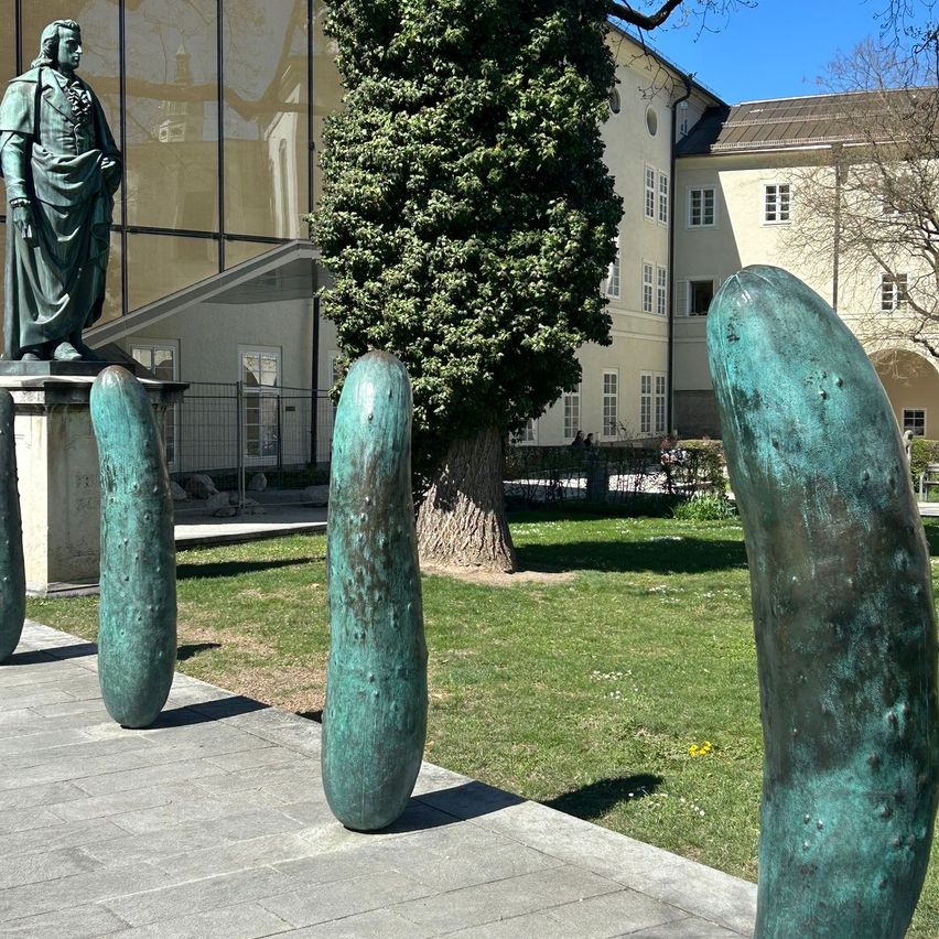 A statue stands on a pedestal in front of a building, with three green pickle sculptures on the pathway. A tree and grassy area are behind the sculptures.