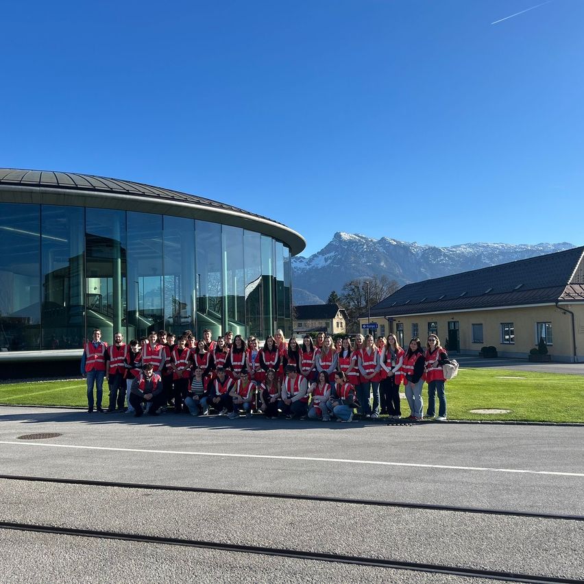 A large group of people in red vests pose for a picture in front of a modern building with glass walls and snowy mountains in the background.