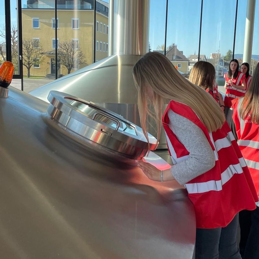 A group of women in safety vests are examining a large metal tank in a brewery. The tank has a silver lid with a handle. They are inside a building with glass walls, and outside there is a building with many windows.