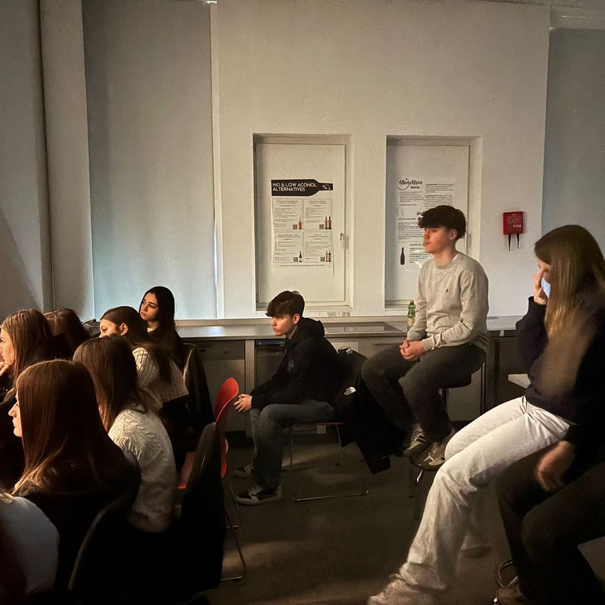 Several students sit in chairs facing a wall with posters. Two males sit on stools at the front, while the others are seated in rows.