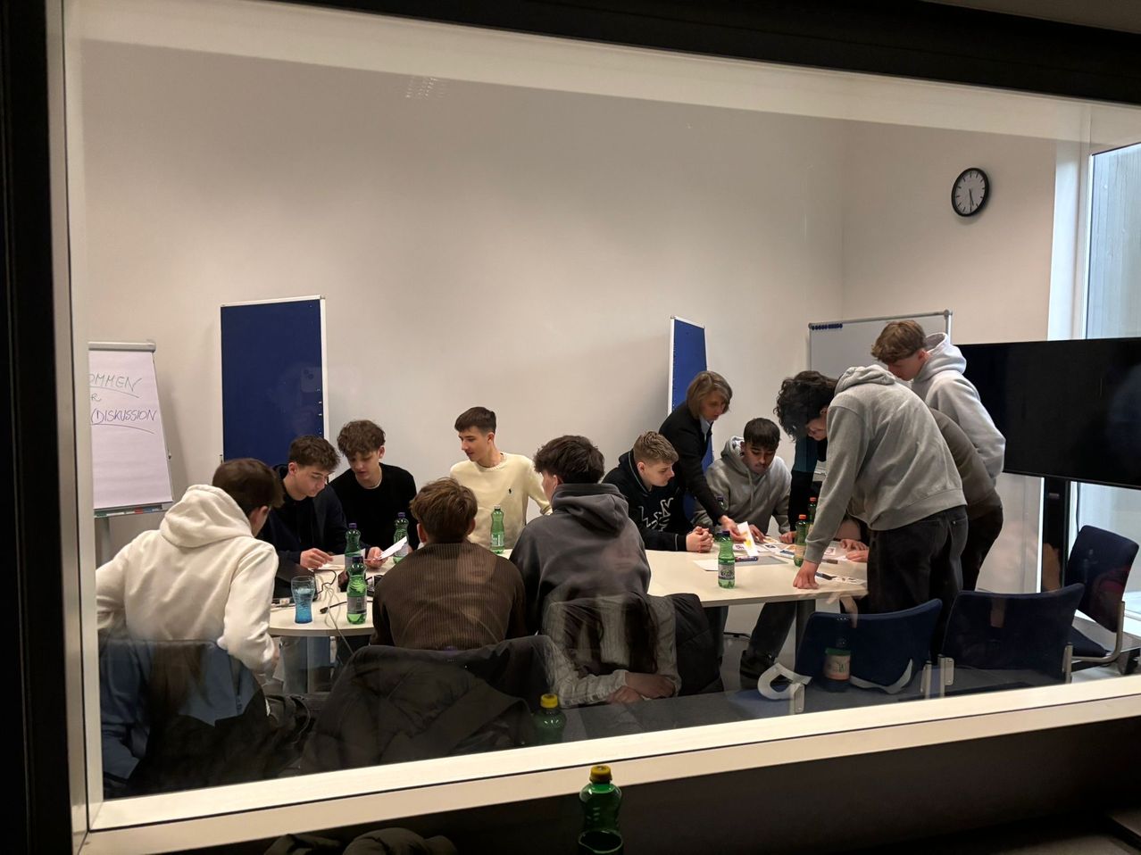 A group of people are sitting around a table in a classroom, possibly working on a project. They are surrounded by bottles and papers.