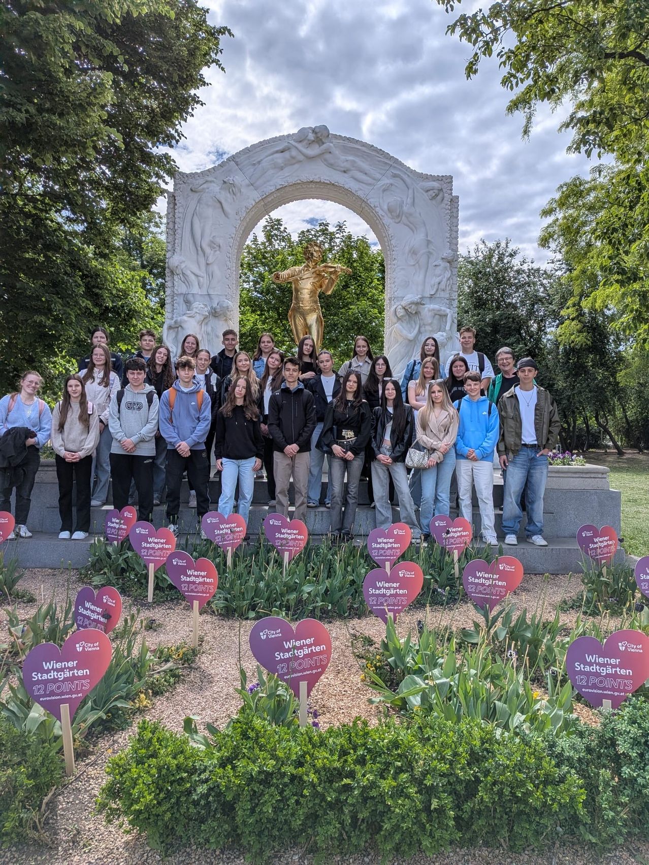 Eine Gruppe von Schülern posiert vor einer goldenen Statue in einem Garten mit herzförmigen Plaketten. Der Himmel ist bewölkt.