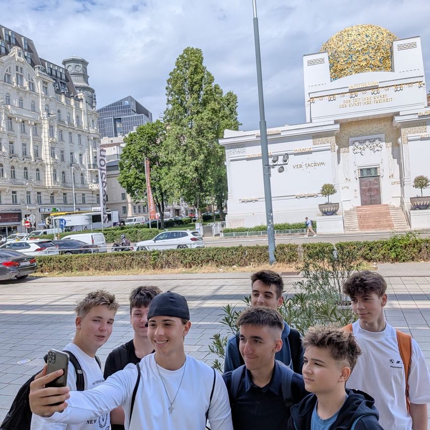 Eine Gruppe junger Männer steht vor einem Gebäude mit einer Kuppel und lächelt. Bäume und geparkte Autos sind im Hintergrund zu sehen.