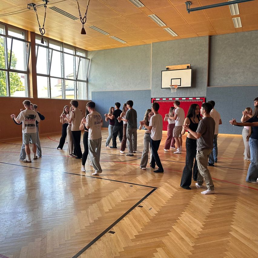 Eine Gruppe von Menschen tanzt in einer Turnhalle mit Holzböden und Fenstern. An der Wand ist ein Basketballkorb angebracht.