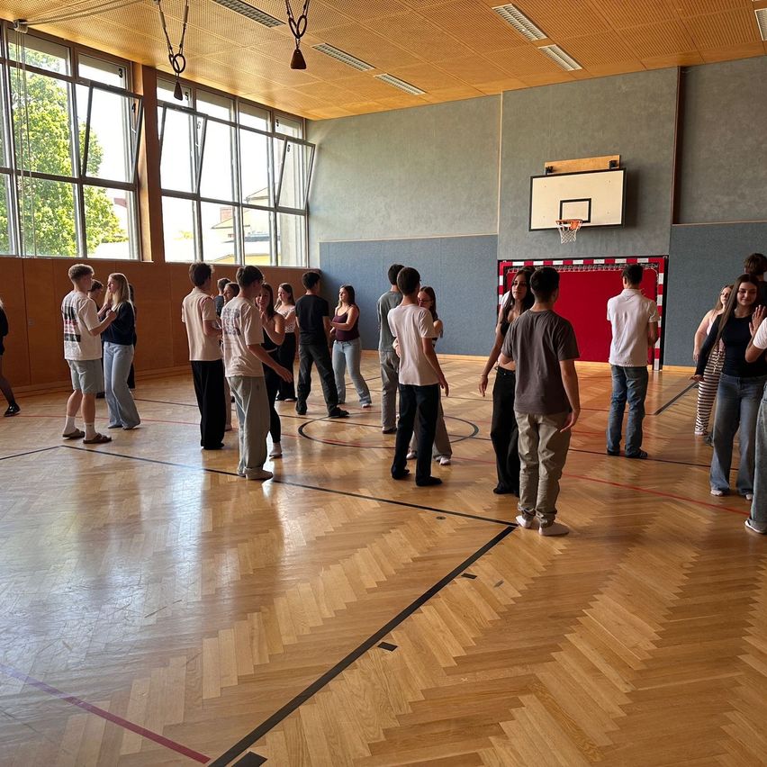 In einer Turnhalle stehen mehrere Menschen in Paaren herum und führen wahrscheinlich ein Gespräch. Die Turnhalle hat Holzböden, einen Basketballkorb und große Fenster.