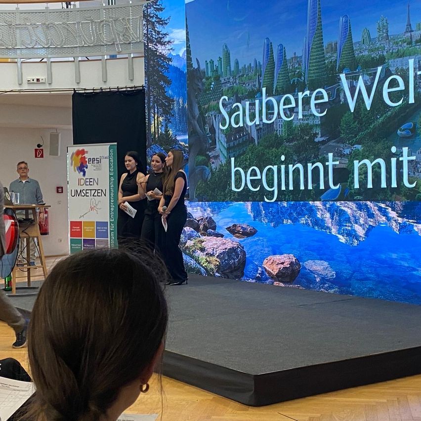 Vier Frauen stehen auf der Bühne vor einer großen Leinwand, die eine saubere Stadt zeigt. Ein Mann steht neben einem Tisch.