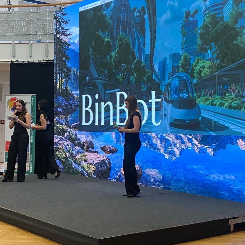 Drei Frauen stehen auf einer Bühne vor einer großen Leinwand, die ein Bild einer futuristischen Stadtlandschaft zeigt. Eine Frau hält ein Mikrofon, eine andere eine Fernbedienung.