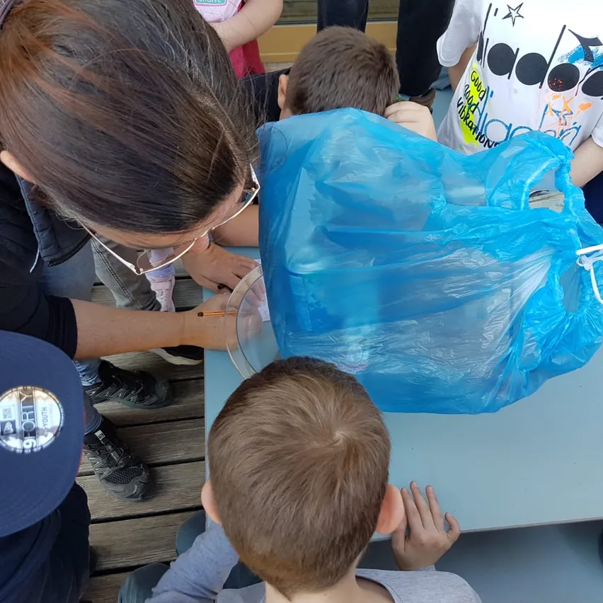 Eine Frau mit Brille und schwarzer Jacke hält einen durchsichtigen Plastikbeutel mit Wasser und gießt den Inhalt in ein Glas. Ein Junge mit blauer Kappe beobachtet sie.