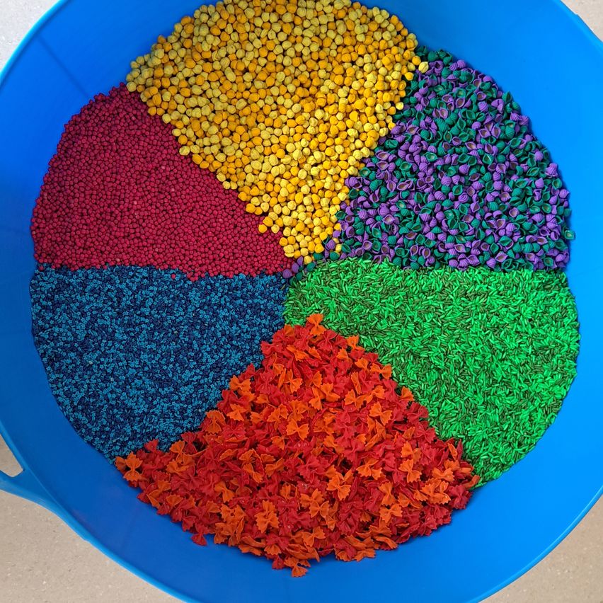 Eine blaue Schüssel ist in sechs Bereiche unterteilt, die mit verschiedenen farbigen Körnern gefüllt sind: gelb, rot, lila, grün, blau und orange.