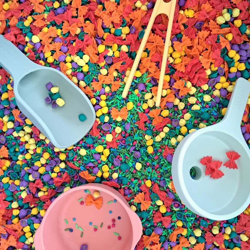 Drei Plastikschaufeln und Chopsticks auf einem Bett aus mehrfarbiger Pasta. Die Schaufeln sind hellblau und rosa, und die Pasta kommt in verschiedenen Farben und Formen.
