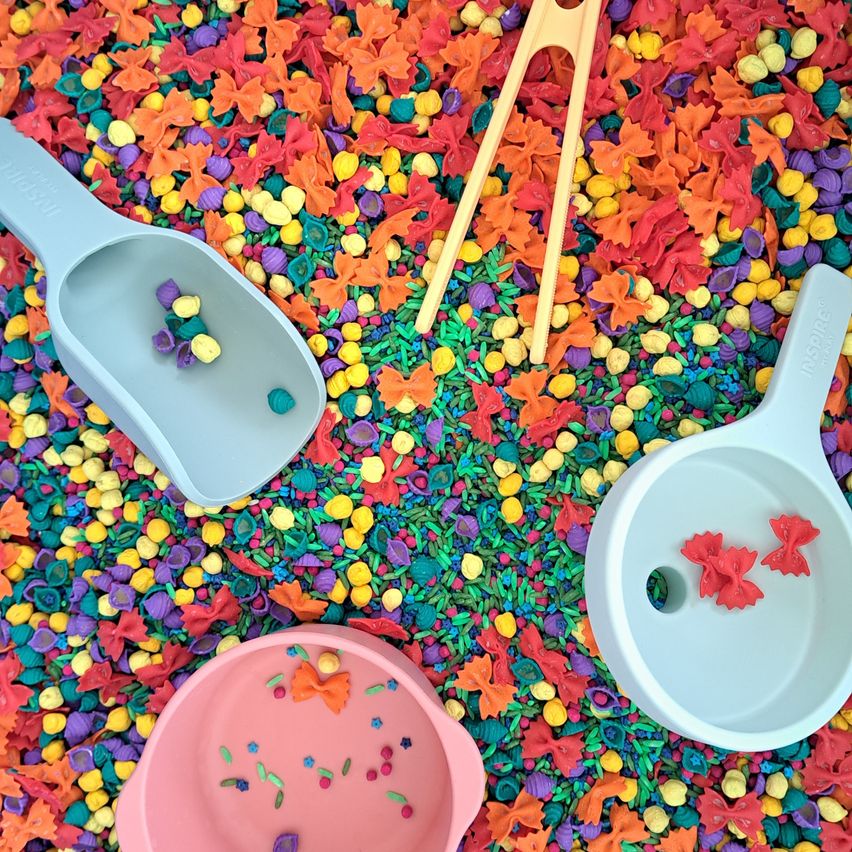 Drei Plastikschaufeln und Chopsticks auf einem Bett aus mehrfarbiger Pasta. Die Schaufeln sind hellblau und rosa, und die Pasta kommt in verschiedenen Farben und Formen.