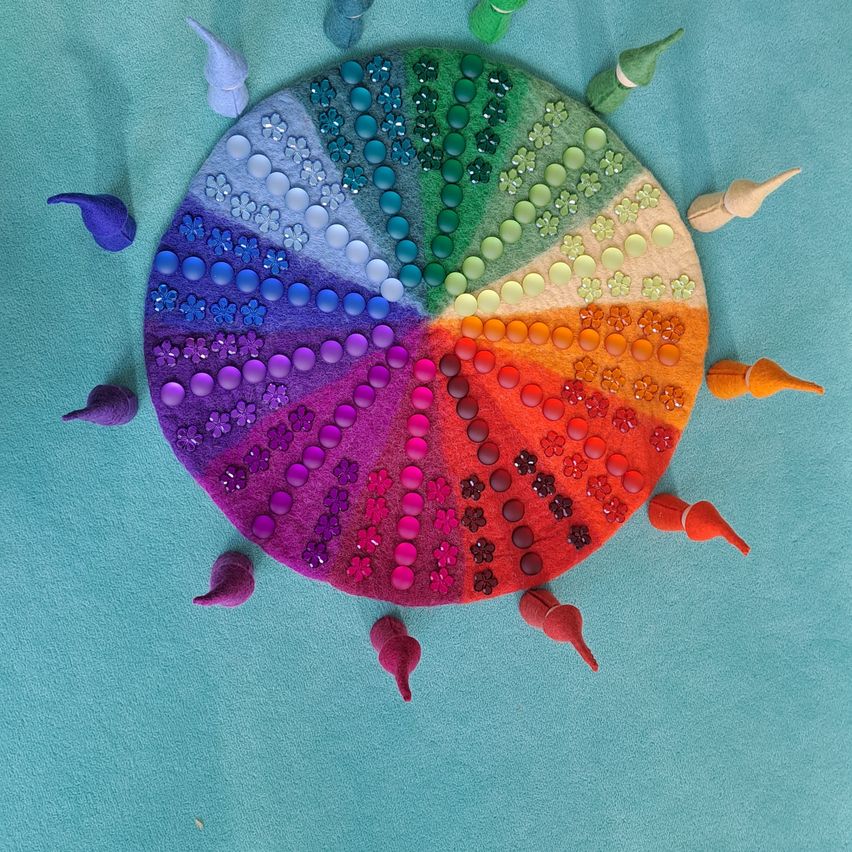Eine bunte kreisförmige Anordnung auf einer blauen Oberfläche mit verschiedenen farbigen Perlen und kleinen Figuren. Die Farben gehen von Blau zu Rot über, dazwischen sind Blumen.