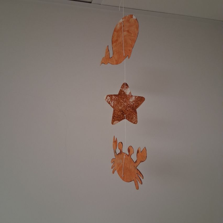 Drei orangefarbene Papierausschnitte in Form eines Fisches, eines Sterns und eines Krebses hängen von der Decke.