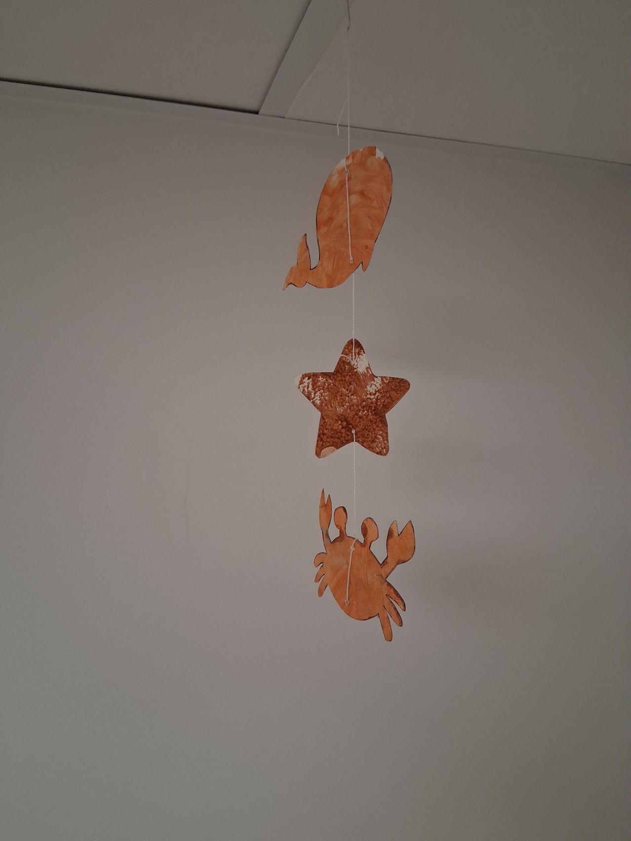 Drei orangefarbene Papierausschnitte in Form eines Fisches, eines Sterns und eines Krebses hängen von der Decke.