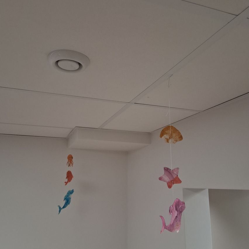 Ein weißer Raum mit zwei dekorativen Mobiles, die Meerestiere darstellen und an der Decke hängen. An der Decke befindet sich auch ein runder Luftauslass.