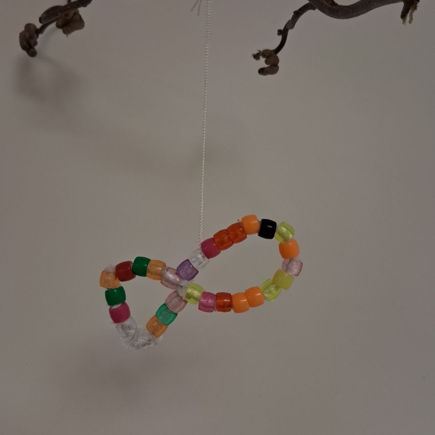 Ein farbenfrohes Perlenfischwerk hängt von einem Ast an einer weißen Wand und zeigt leuchtende Farbtöne von Orange, Grün und Rosa.