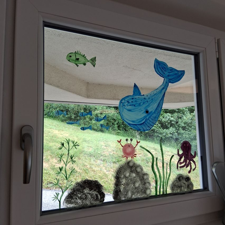 Ein Fenster mit Fischaufkleber, das einen Wal, Fische, Krabben und einen Oktopus zeigt. Darunter sind Felsen, Seegras und ein Grasfeld sichtbar.