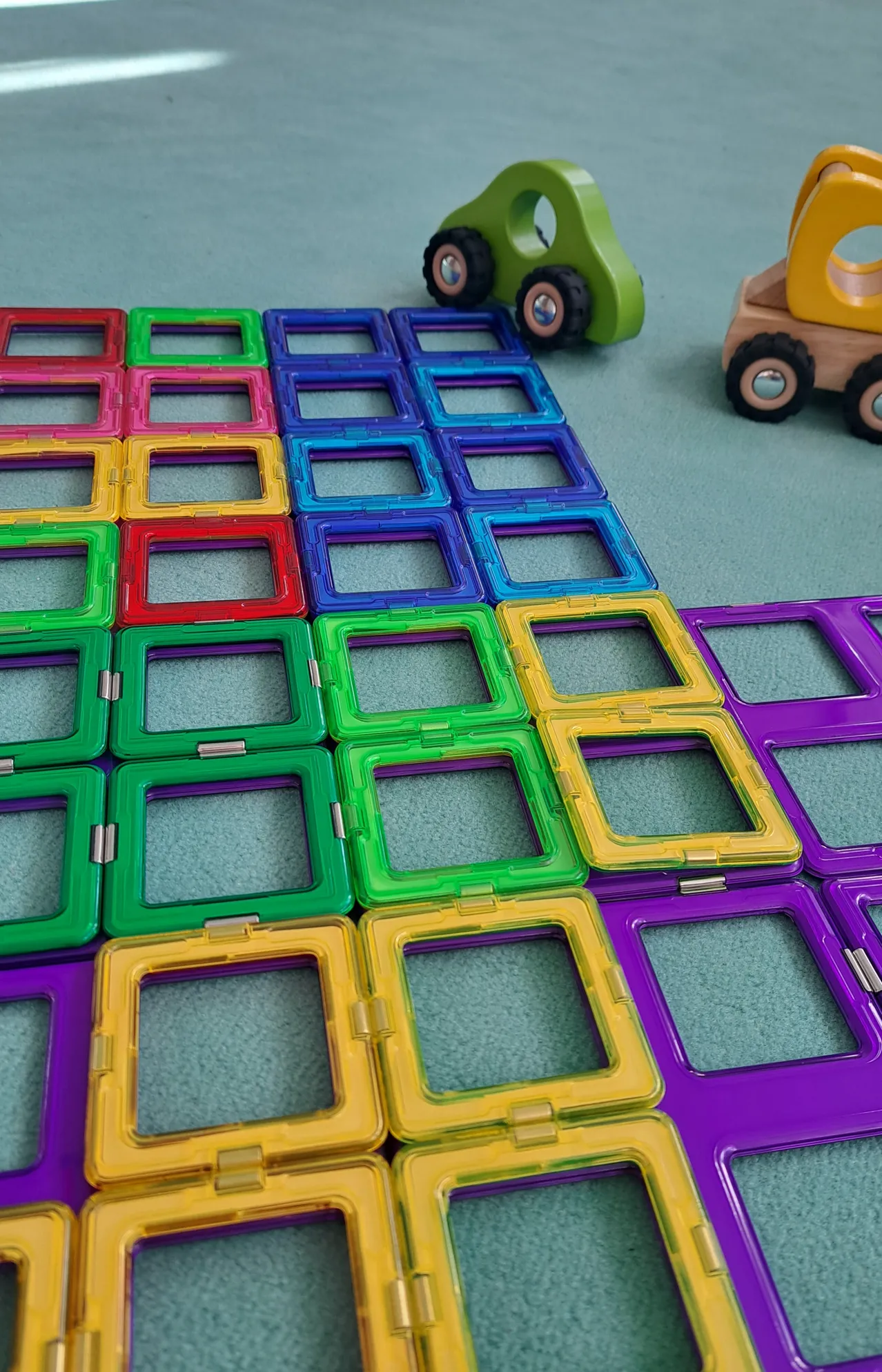 Eine Sammlung bunter, magnetischer Spielsteine auf einem Teppichboden, angeordnet in einem Muster mit Spielzeugfahrzeugen in der Nähe.