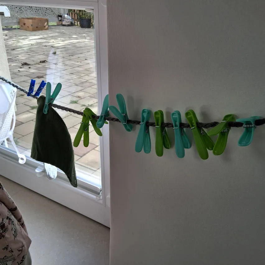 Eine Person hängt Kleidung an einer Schnur mit grünen Wäscheklammern an einer Wand.